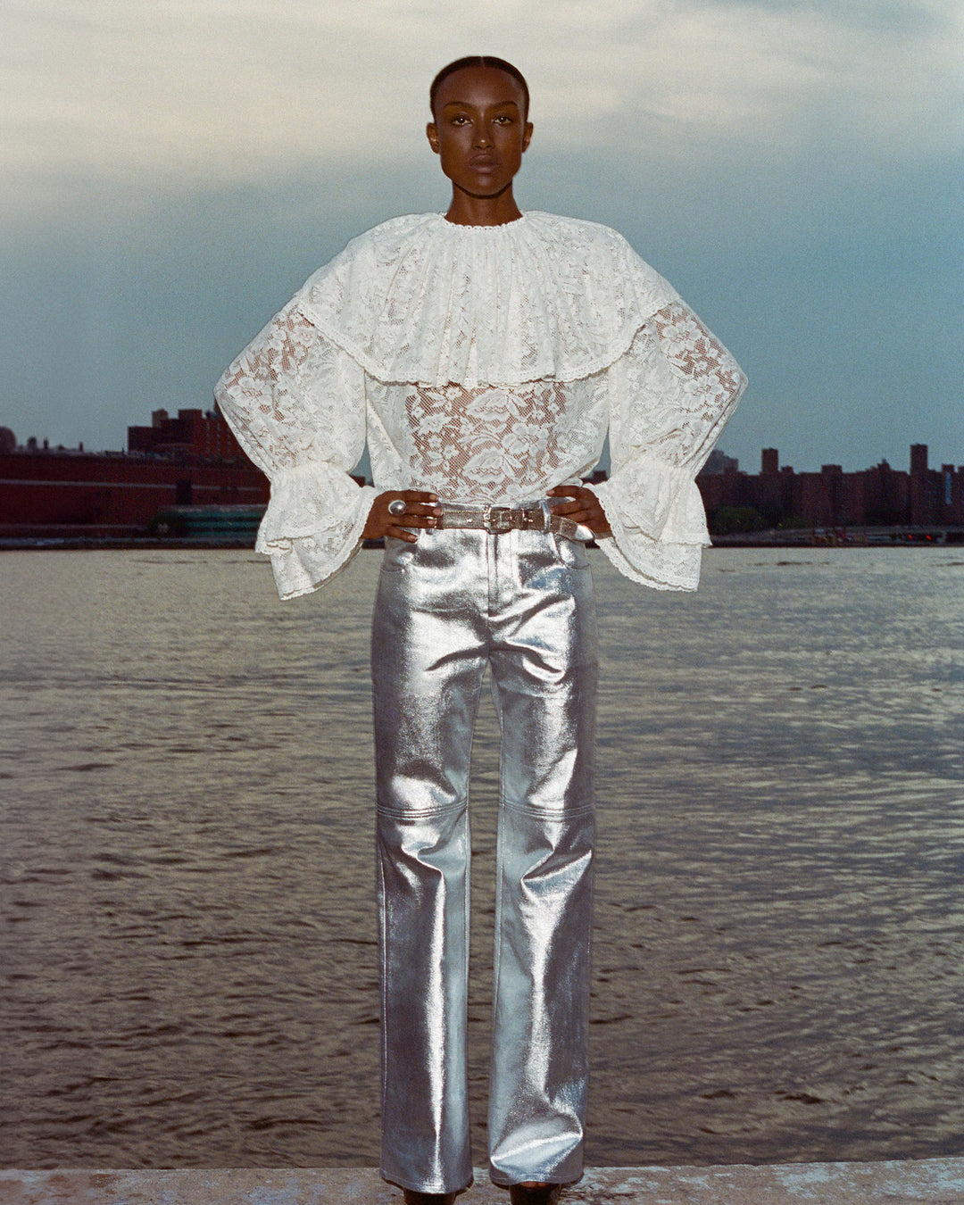 A person wearing the Eloise Magnolia Lace Top, which has a vintage floral lace design, and shiny silver pants stands by the waterfront, framed by buildings in the background.