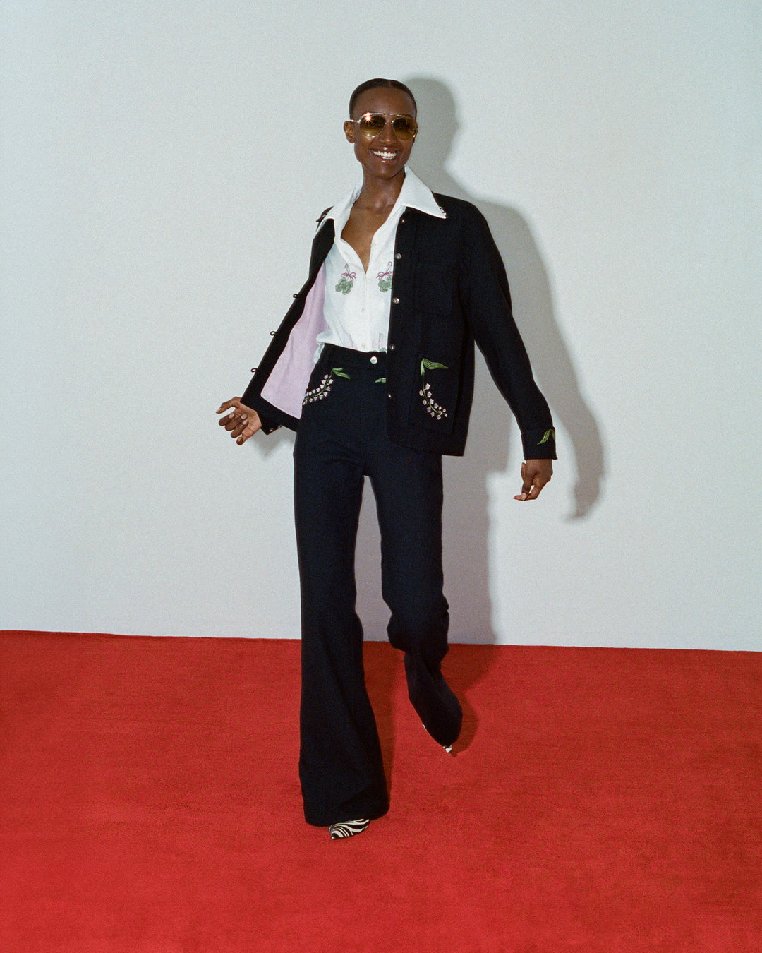 Wearing a pair of sunglasses, a black jacket, and the stylish Stevie Lucky Clover Shirt, an individual strolls confidently on the red carpet in black flared pants featuring hand-embroidered details.