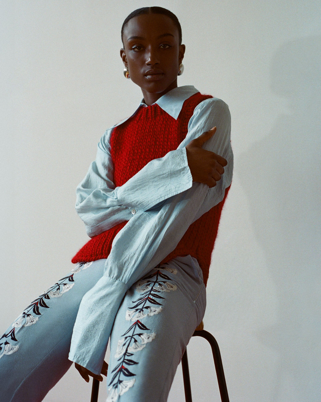 A person is sitting on a stool, wearing the Quentin Cardinal Knit Top over a blue shirt, paired with embroidered jeans, in front of a plain background.