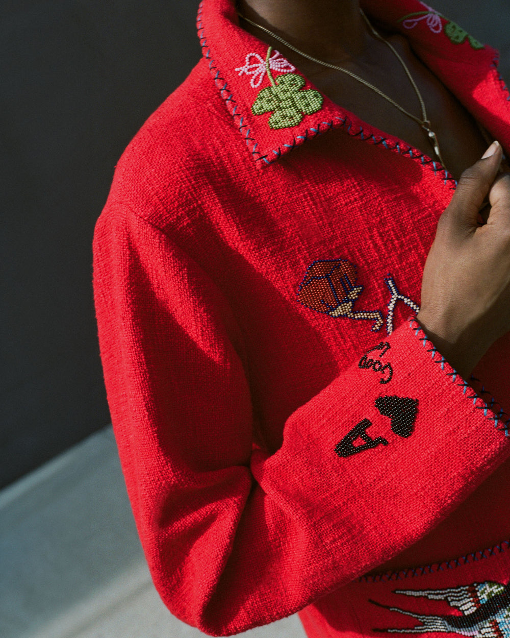 A person wearing the Francis Crimson Tattoo Jacket, featuring a vintage design with intricate red embroidery that includes playing card symbols and a balloon, showcases the detailed craftsmanship. The focus is on the torso and hand adjusting the collar of this hand-beaded masterpiece.