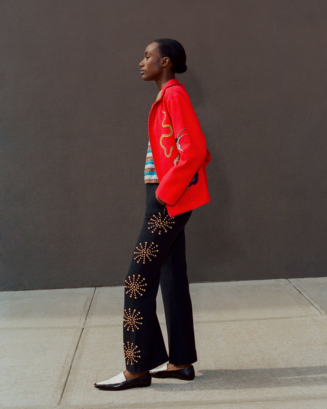 A person stands against a plain background wearing a Francis Crimson Tattoo Jacket with a vintage feel, paired with a colorful top and black pants featuring star patterns. Their stylish look is completed with loafers.