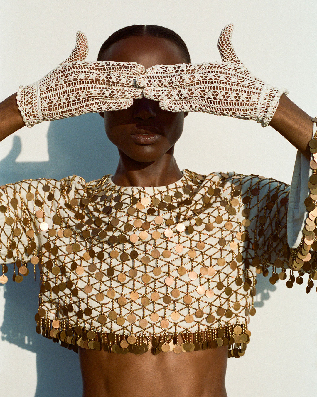Wearing the Nina Whitegold Coin Top and white crochet gloves, a person covers their eyes with their hands against a plain background.