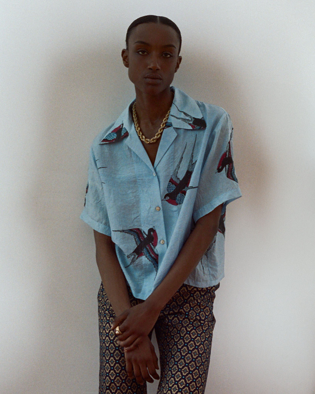 Against a plain background, a person wears the Stevie Swallow Silk Shirt, light blue with bird prints like a vintage Hawaiian style. They pair it with patterned pants, sport short hair, and accessorize with a gold chain necklace.