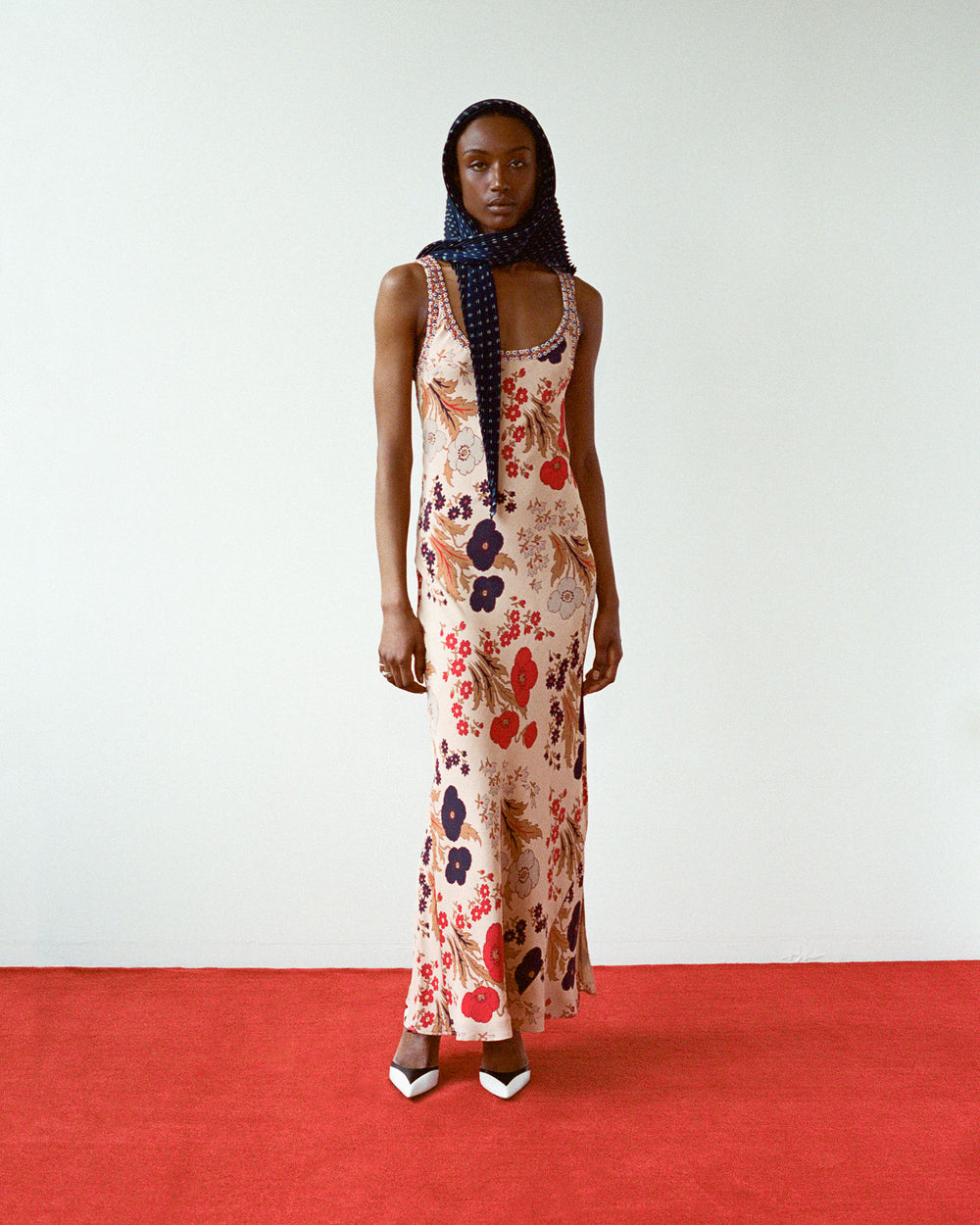 Standing on a red carpeted floor against a white wall, the person wears an Adele Vintage Poppy Dress featuring red, blue, and beige floral patterns. A dark scarf is tied around their head, and they finish the look with black and white pointed shoes.