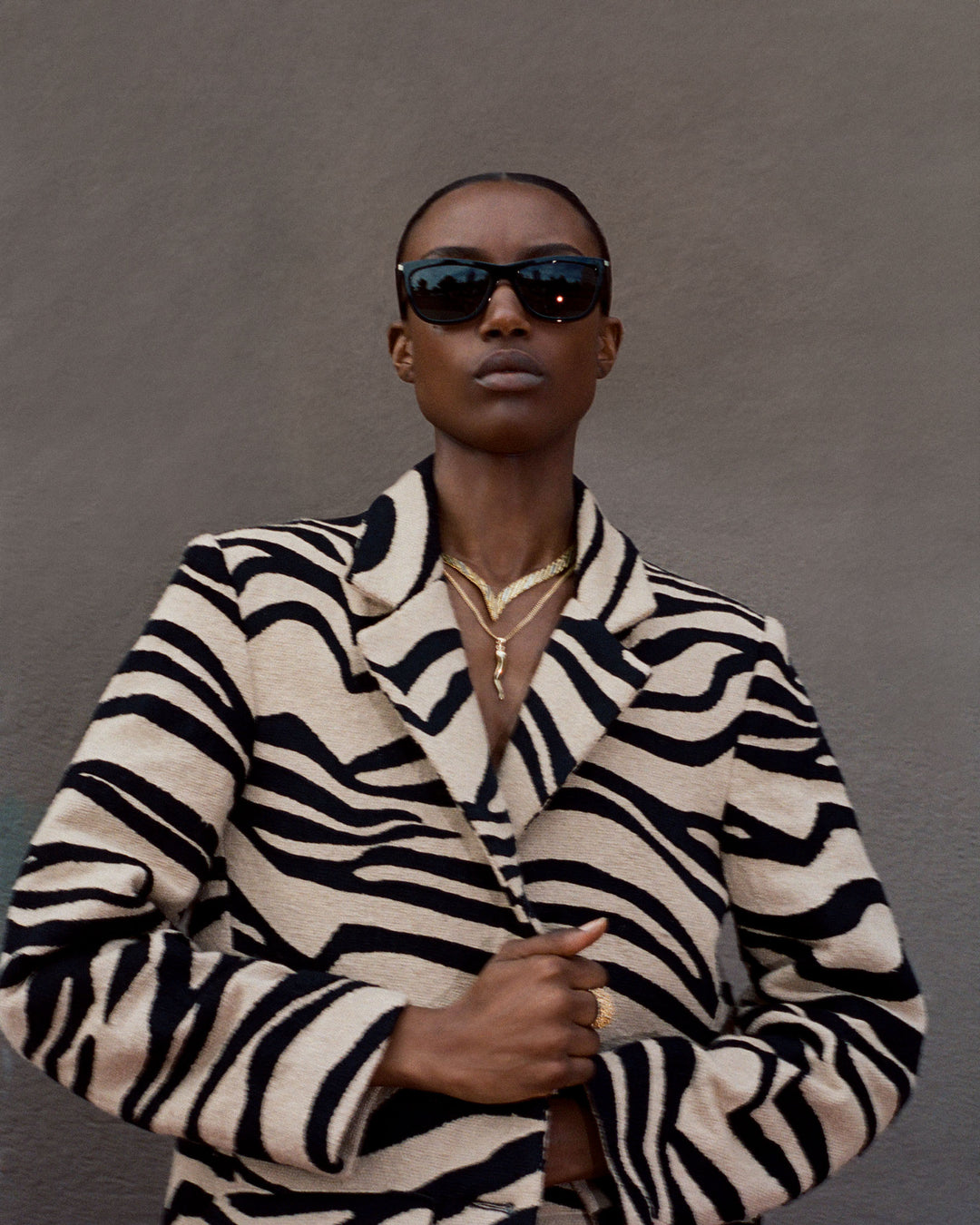 Dressed in the Captain Zebra Mirage Coat, chic sunglasses, and elegant gold necklaces, a person stands confidently against a muted gray background.