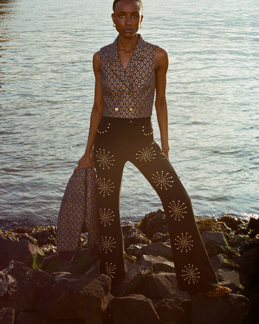 A person stands confidently on rocks by the water, wearing the Leo Graceland Vest and flared black pants adorned with star-like designs. Their matching jacket features gleaming St. Christopher buttons, while the setting sun casts a warm glow on the scene.