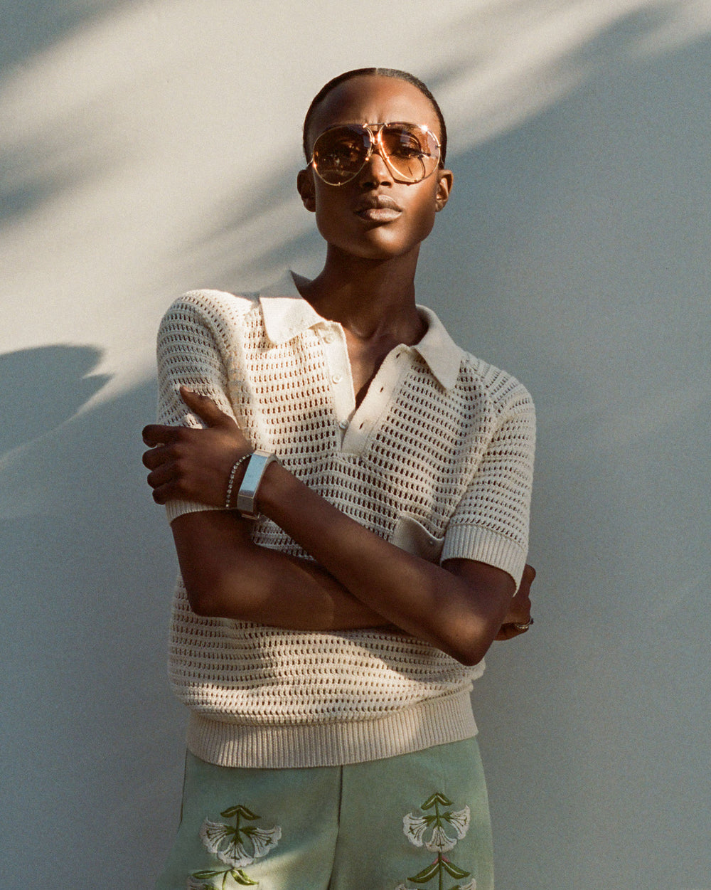 Clad in the Lessi Oyster Crochet Top and vintage-inspired sunglasses, a person stands with arms crossed, channeling a Katharine Hepburn vibe. Shadows play on the wall behind, enhancing the classic charm.