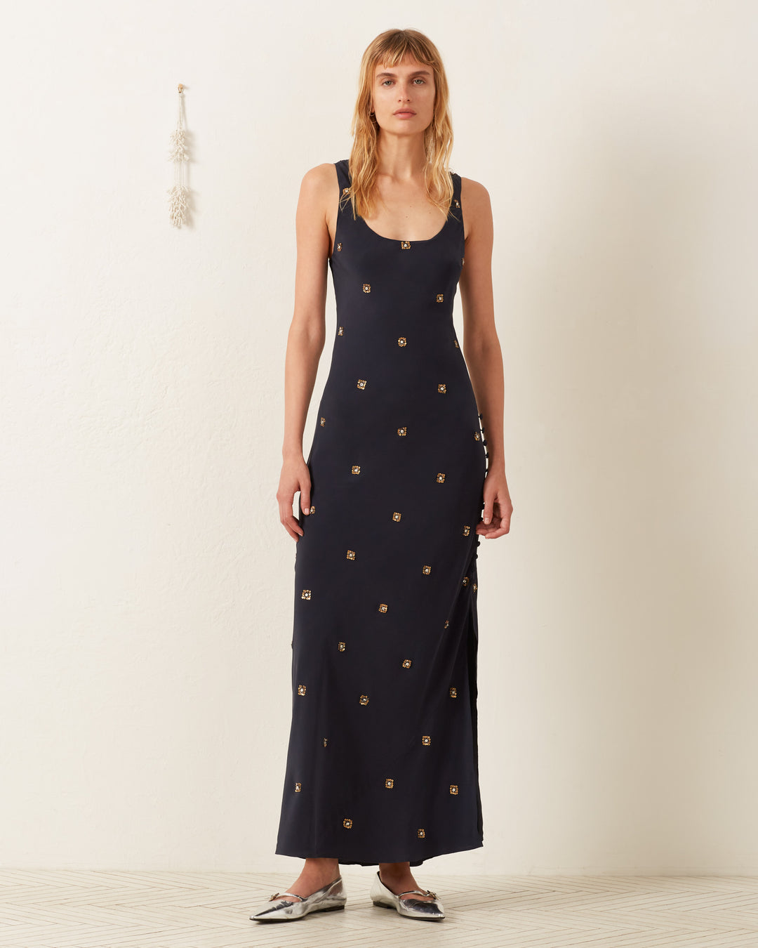 Against a plain white background, someone models the Adele Casino Night Dress, a black sleeveless slip with gold geometric designs. They have shoulder-length hair and wear silver pointed shoes. A small decorative wall hanging is on the left.