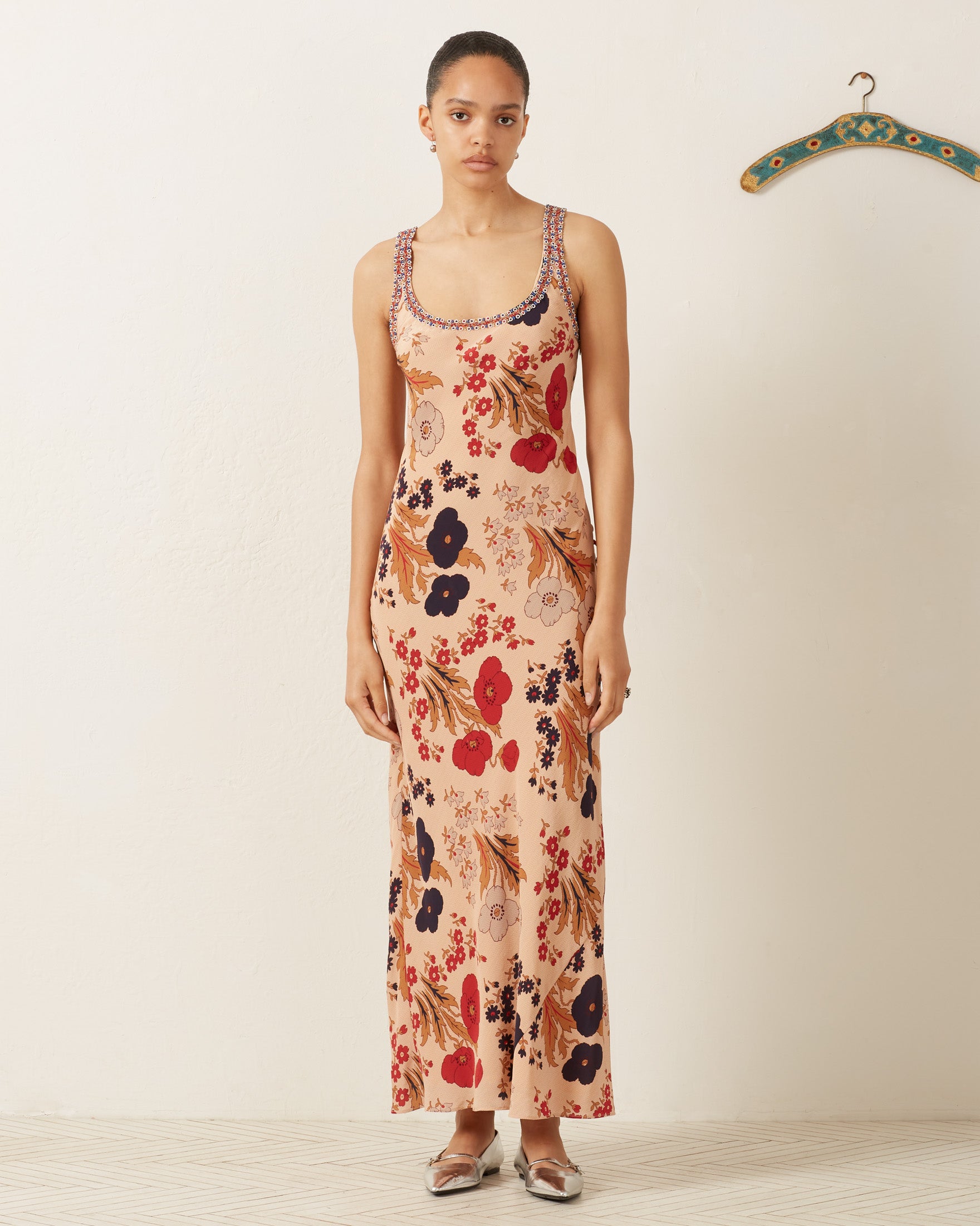 In front of a white wall, a person wears the Adele Vintage Poppy Dress, adorned with red, black, and brown floral patterns, paired with silver flats. A decorative hanger can be seen on the right.