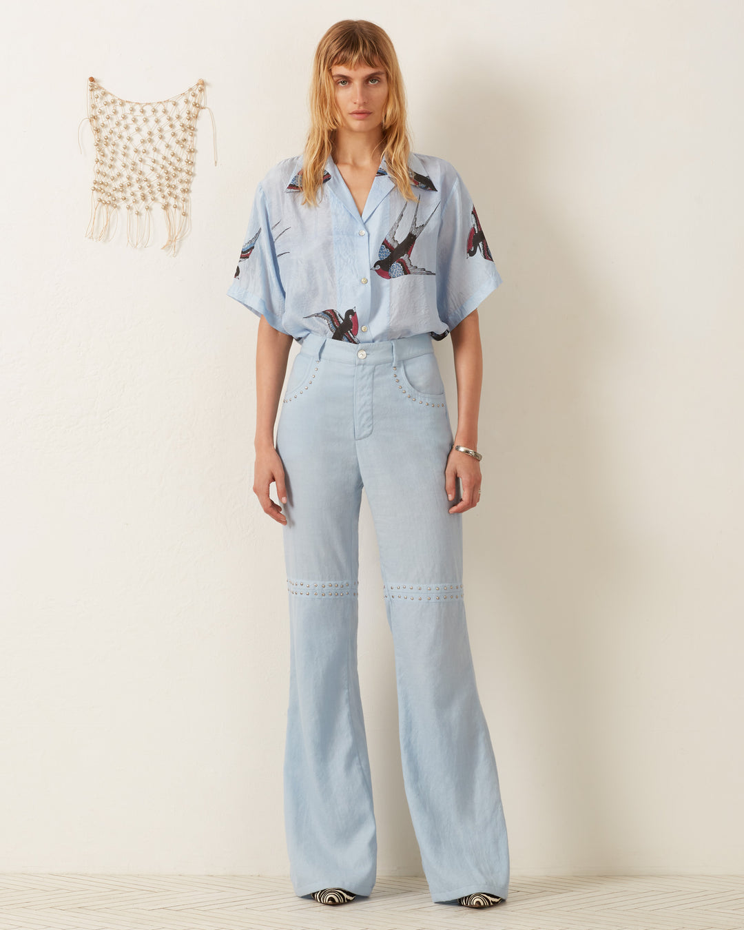 A person with long hair stands against a plain white wall wearing the Stevie Swallow Silk Shirt, a light blue button-up featuring hand-block printed bird designs, paired with high-waisted, light blue flared jeans. A small fringed macramé decoration hangs on the wall.