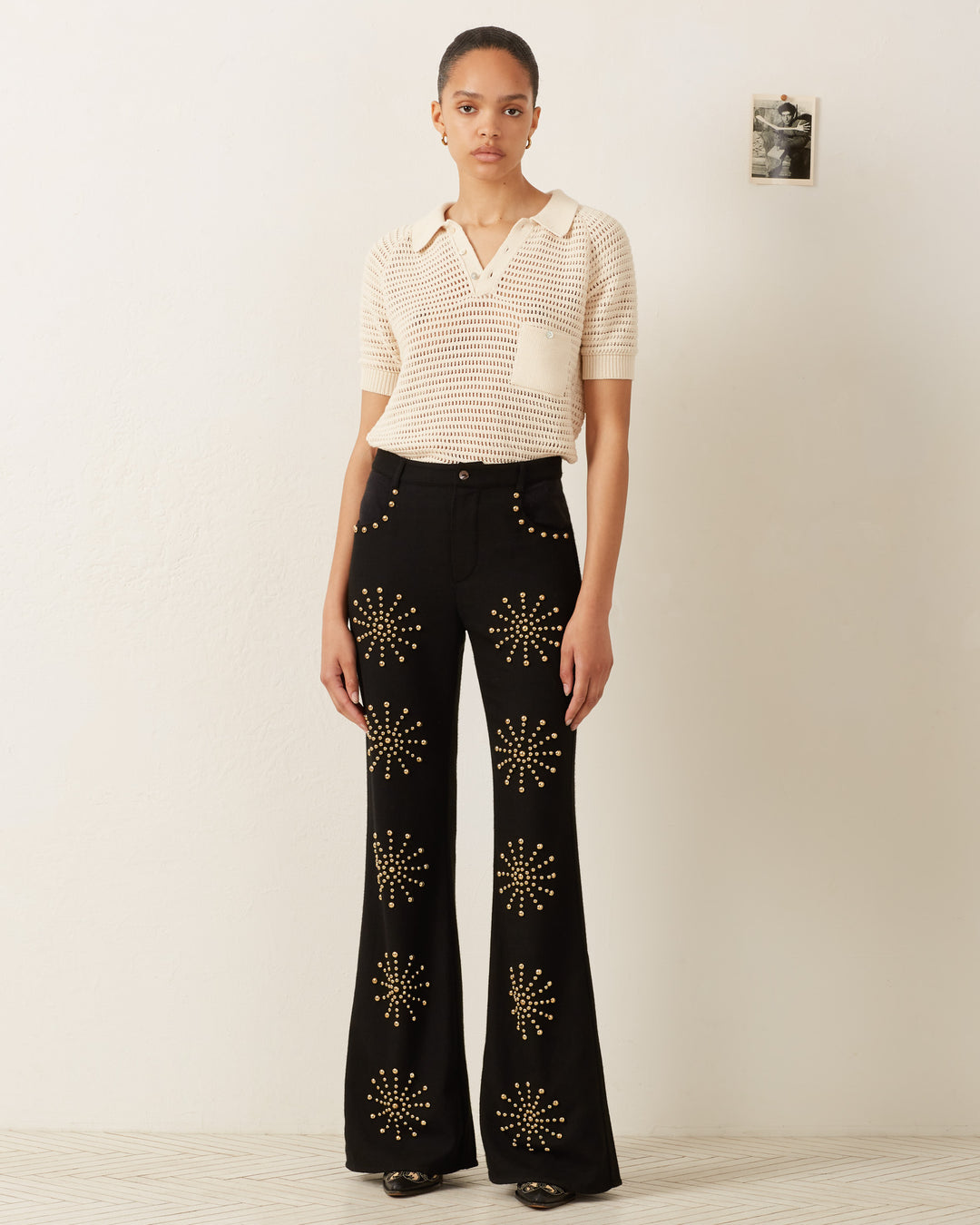 A woman stands against a plain wall, exuding Katharine Hepburn's style with vintage flair. She wears the Lessi Oyster Crochet Top and black flare pants with star patterns, effortlessly blending modern chic with classic elegance.