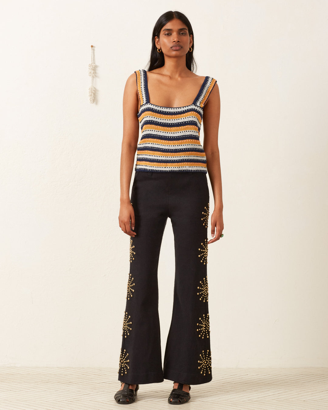 A person with long dark hair wears the Mia Mirage Crochet Top and black flared pants with patterns. They stand against a plain wall featuring a small decorative hanging, complemented by a light-colored textured floor that adds subtle charm to the scene.