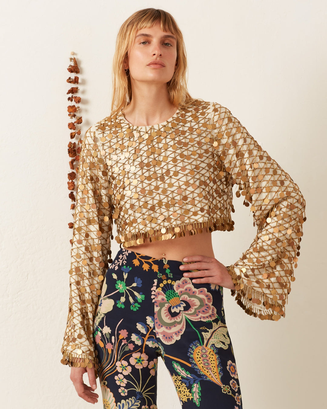 Wearing the Nina Whitegold Coin Top, a hand-embellished cropped piece with gold sequins and bell sleeves, paired with high-waisted dark floral pants, this person stands against a plain wall.