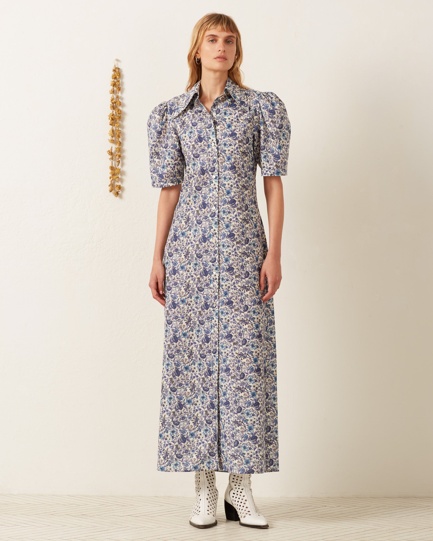 A person stands against a plain wall wearing the elegant Deneuve Luna Flower Dress with puffed sleeves and white ankle boots. A small decorative item hangs on the wall beside them.