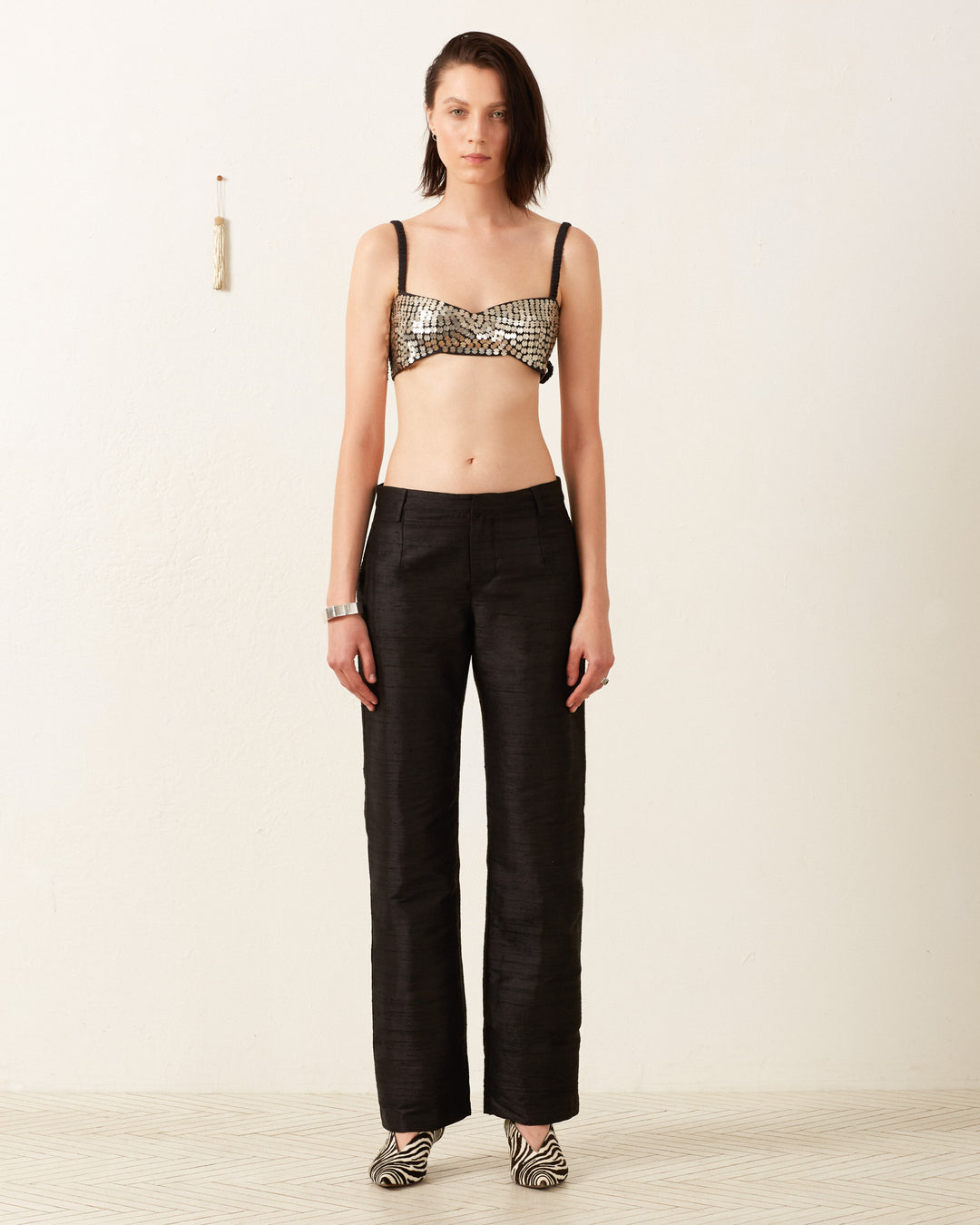 A person stands against a plain white wall, wearing a metallic checkered bralette, Elinor Blackjack Silk Pant, and zebra-patterned pointed shoes. Their straight hair frames their face as they look directly at the camera. A small hanging decor is visible on the wall.