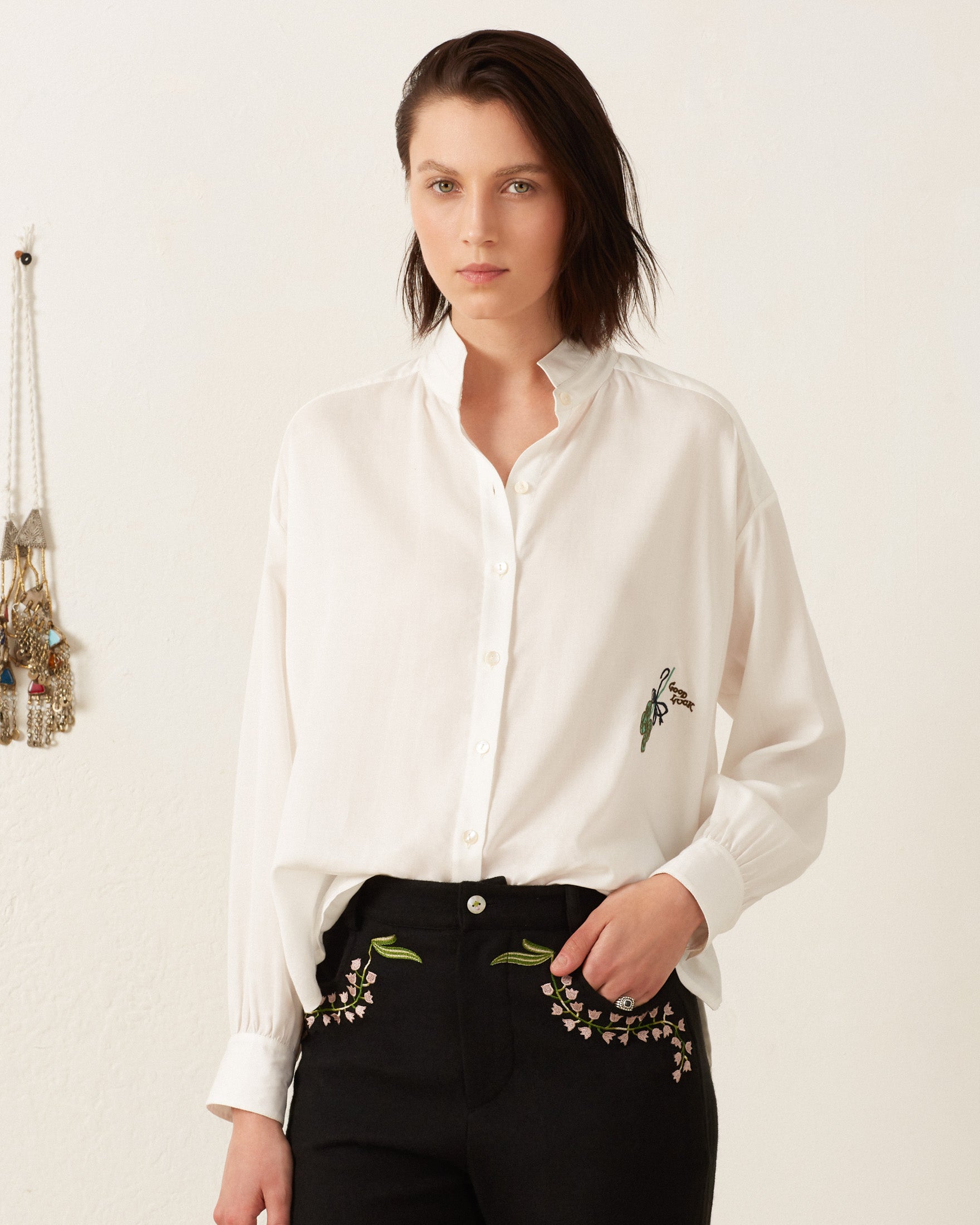 A person in a Kiki Lucky Clover Shirt, featuring hand-embroidered details, and black pants stands against a light-colored background decorated with hanging items.