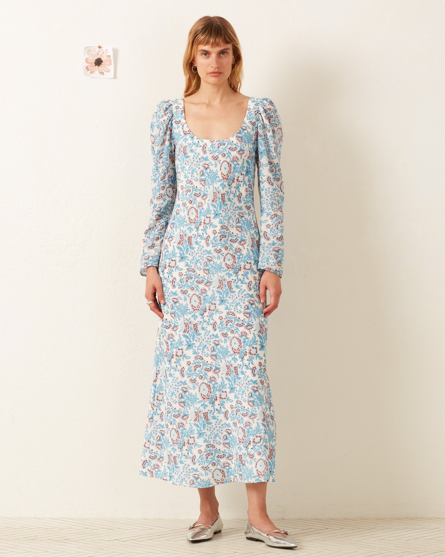 A woman stands against a white wall wearing the Paloma Blue Garden Dress, a blue and pink maxi crafted from a soft cotton-silk blend, paired with silver flat shoes.