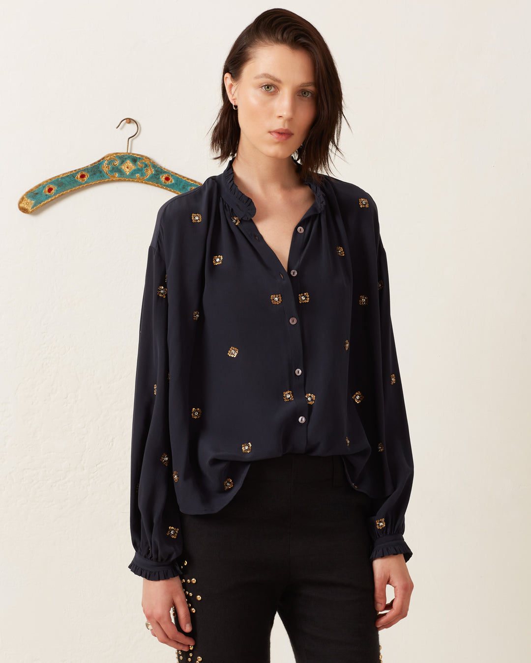 A person with short dark hair stands against a plain wall wearing the Poet Casino Night Blouse, a vintage-inspired black button-up shirt with a patterned design. A colorful clothes hanger is visible on the wall behind them.