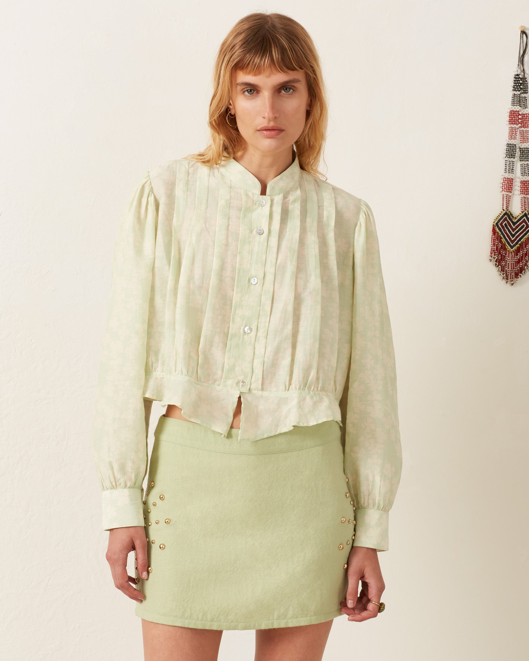 A woman stands against a plain white background, wearing the Ryland Celadon Clover Shirt, a Victorian silhouette with pleats, and a short elegant skirt featuring button details. Her shoulder-length hair frames her neutral expression. A beaded decoration subtly complements the scene.