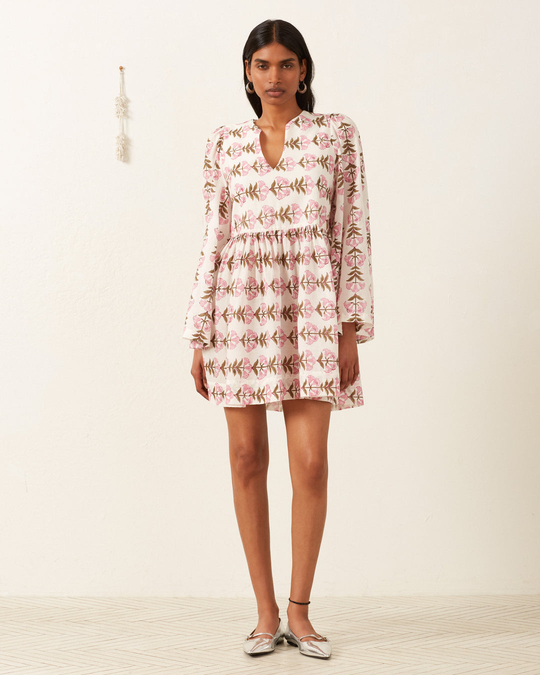 Standing against an off-white wall, a person wears the Silvie Trumpet Lily Dress in soft pink and green floral print. Silver shoes complete the look, with a small decorative item resting on the textured, light-colored floor.