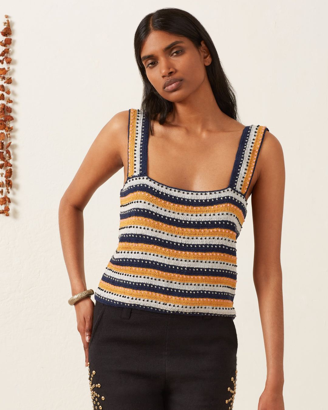 Against a light backdrop, a person with dark hair poses with one hand on their hip, wearing the sleeveless Mia Mirage Crochet Top in navy, gold, and white stripes. To the left, playful strands of small multi-colored beads enhance the scene.