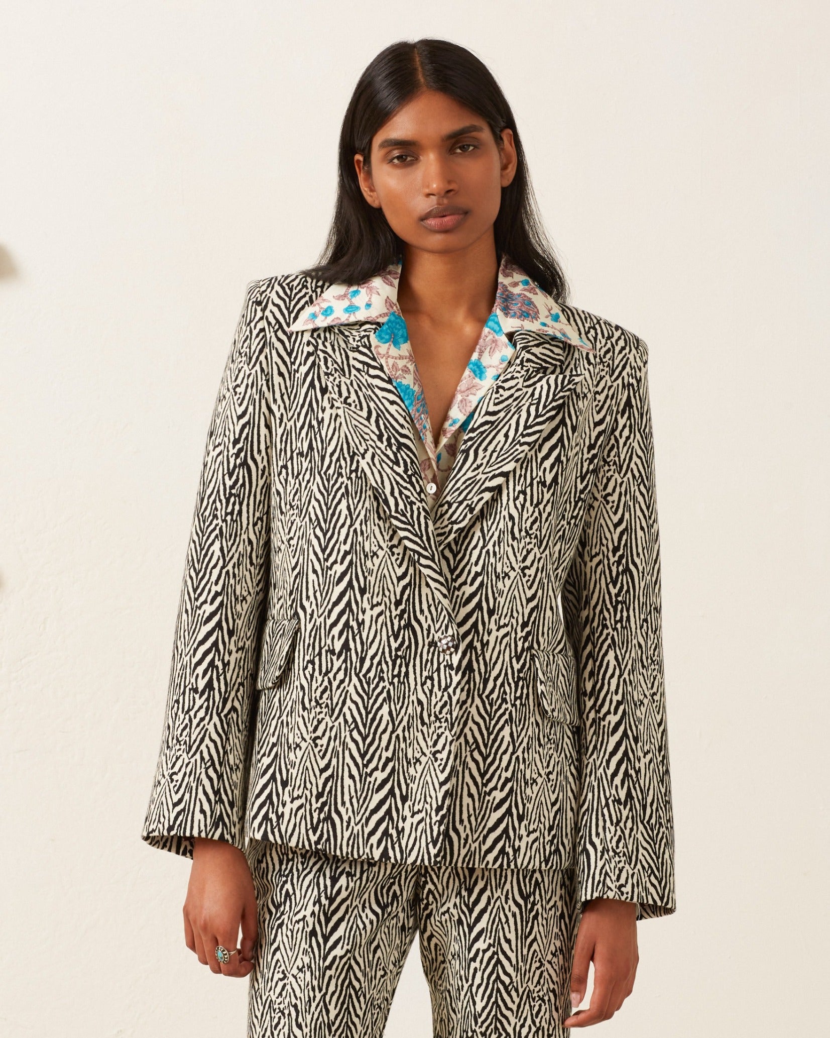 A person with long dark hair poses in front of a plain backdrop, wearing the Dree Zebra Blazer made of virgin wool. The black and white blazer has a colorful floral collar, peak lapels, and they stand with hands relaxed at their sides.