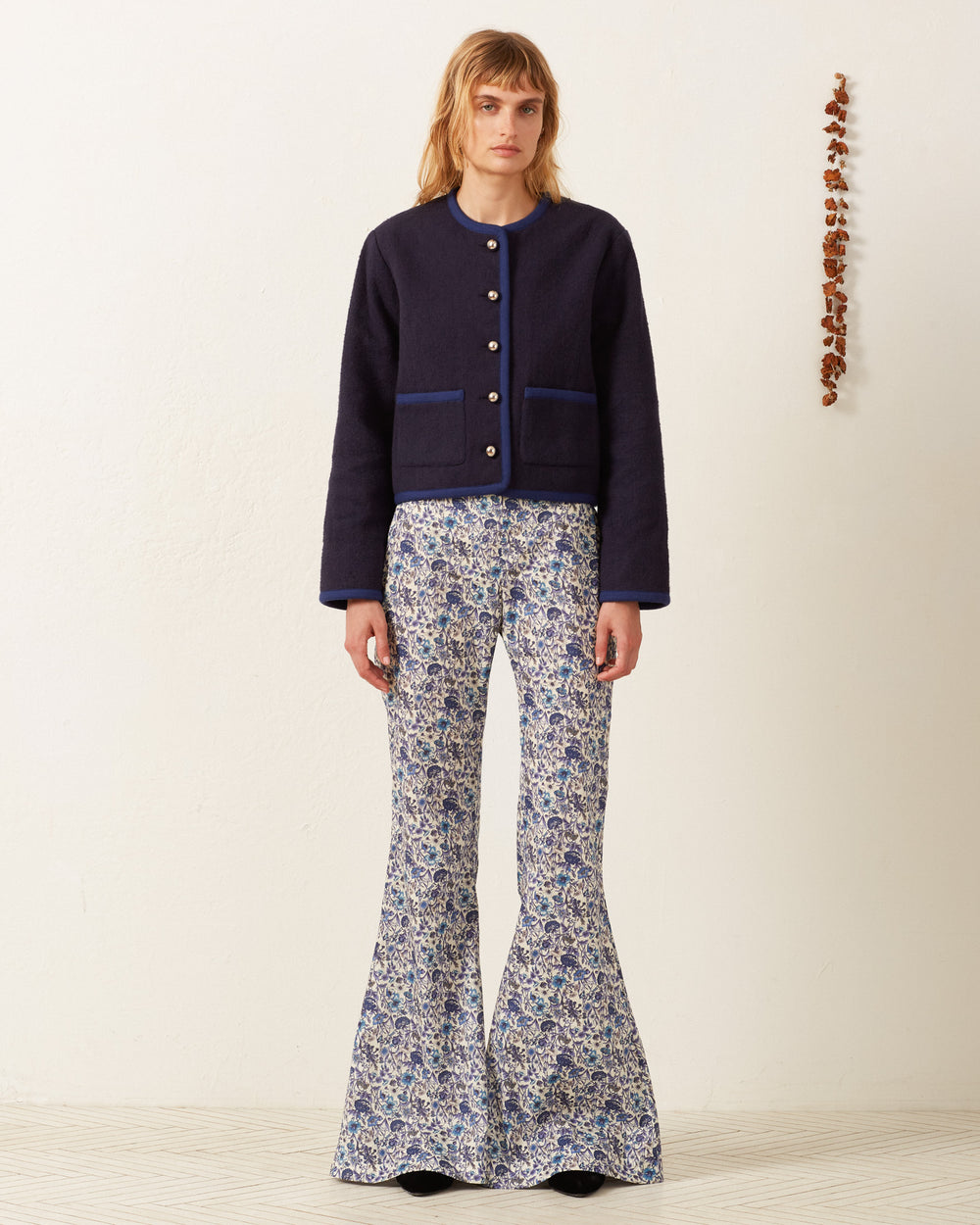 A person stands against a white wall wearing the Rowan Midnight Swallow Jacket, featuring large buttons, with flared pants adorned with blue and white floral designs. Beside them, a thin vertical arrangement of small brown objects showcases an artisan's hand in each delicate detail.