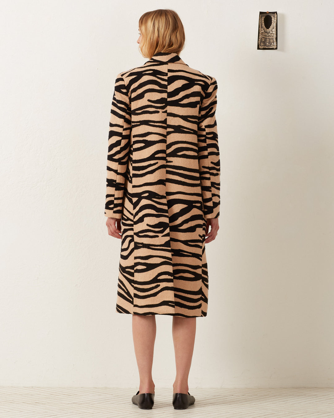 A person wearing the Captain Zebra Mirage Coat, a long, beige and black animal print design, stands with their back turned in a minimalistic room featuring a tasteful wall decoration.