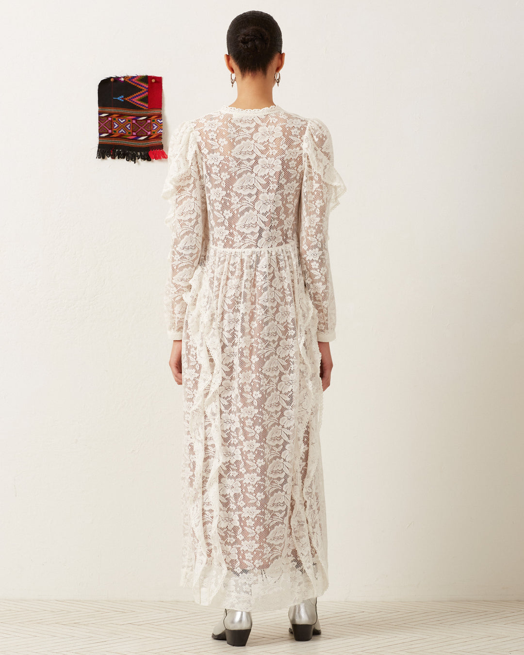 A person in the Lina Magnolia Lace Dress, an ivory maxi with ruffled trim, stands against a plain white wall. To the left, a colorful patterned textile hangs. The individual's back faces the camera, completing their vintage bridal look with silver shoes.