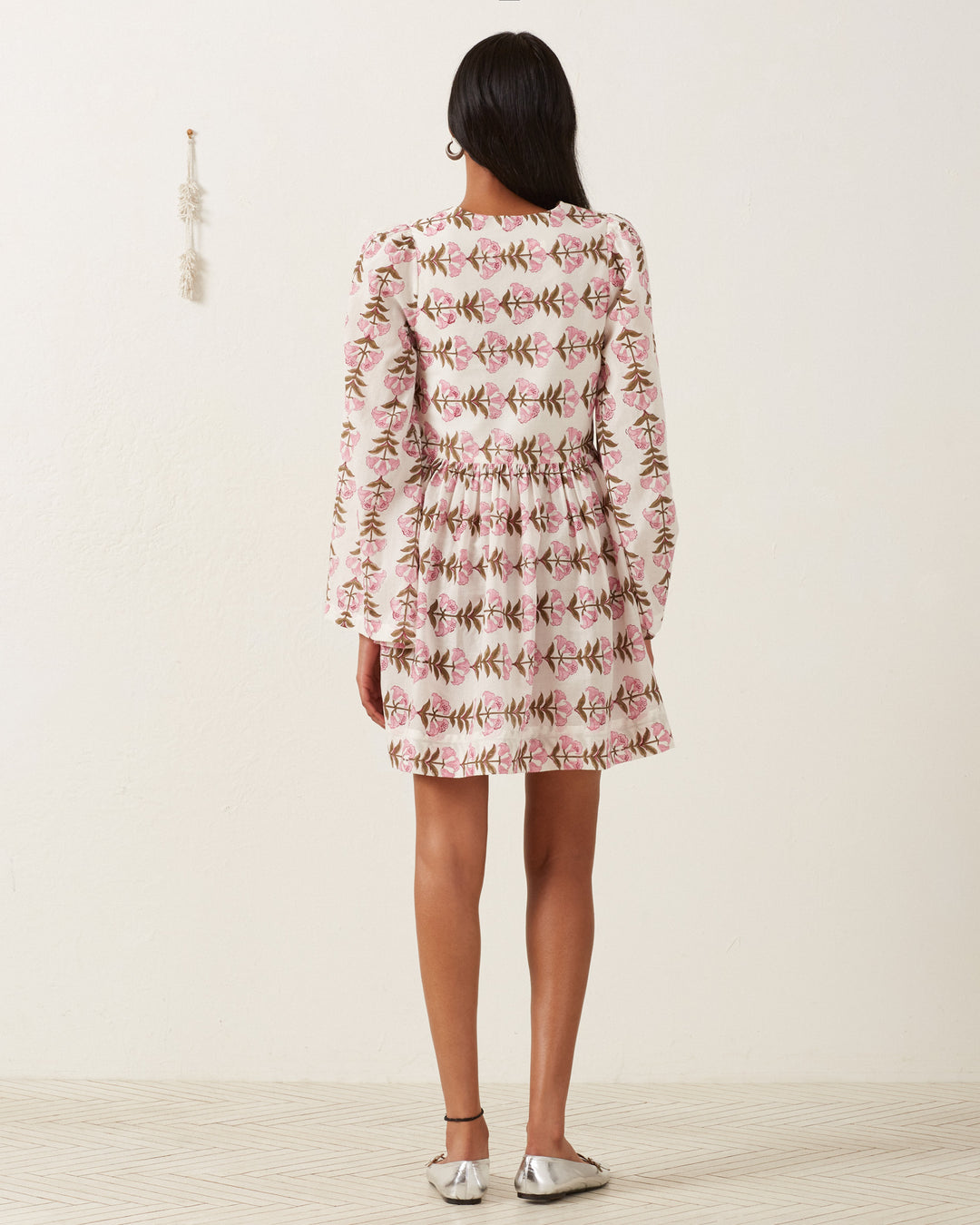 A person with long dark hair stands facing away in a Silvie Trumpet Lily Dress featuring hand-block printed, floral patterns in soft pink and green. The knee-length cotton voile flows gently, complemented by metallic silver flats on a beige textured floor.