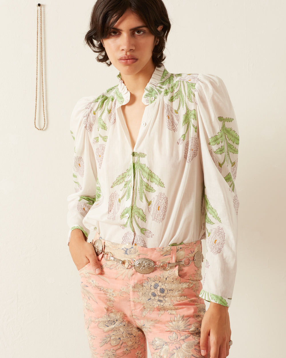 A person stands against a plain backdrop wearing the Annabel Moonflower Shirt, featuring green and pink hand block printed floral designs, paired with matching pink floral pants. They have one hand in their pocket and a neutral expression. A thin chain hangs on the wall behind them.