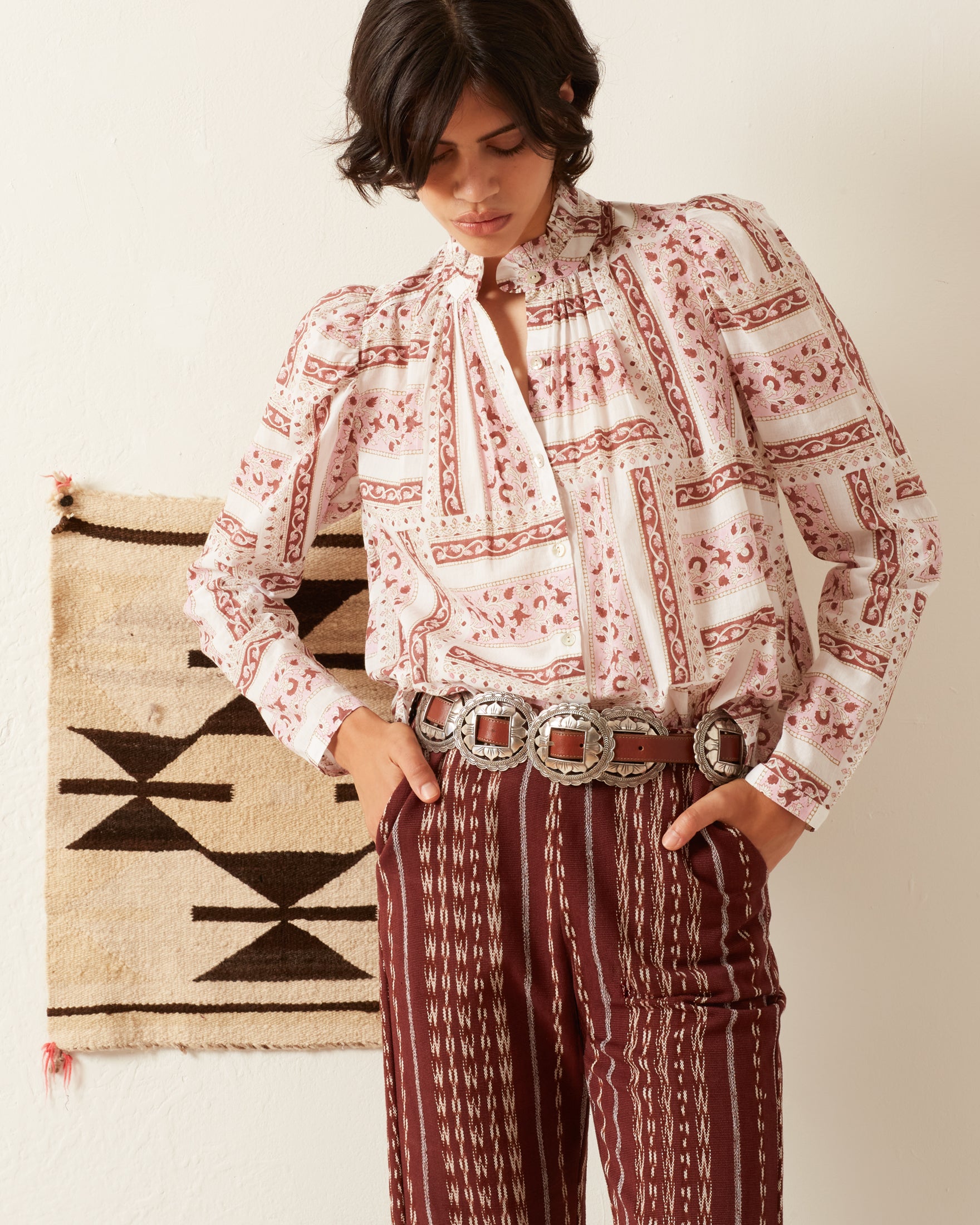 A person with short dark hair wears the Annabel Rosé Shirt, which features a pattern and puffed sleeves, along with high-waisted patterned pants enhanced by a decorative belt. They stand in front of a wall adorned with a small woven tapestry, their head tilted slightly downward.