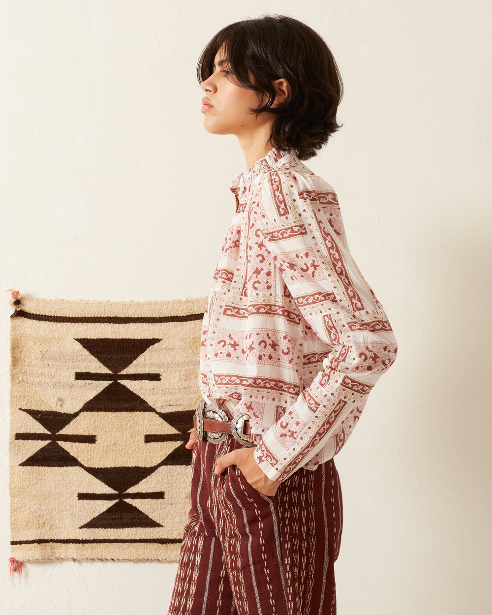 A person with short, dark hair stands sideways, dressed in the Annabel Rosé Shirt with a subtle ruffled collar and long sleeves, paired with pants. A geometric-patterned textile hangs on the off-white wall behind them.