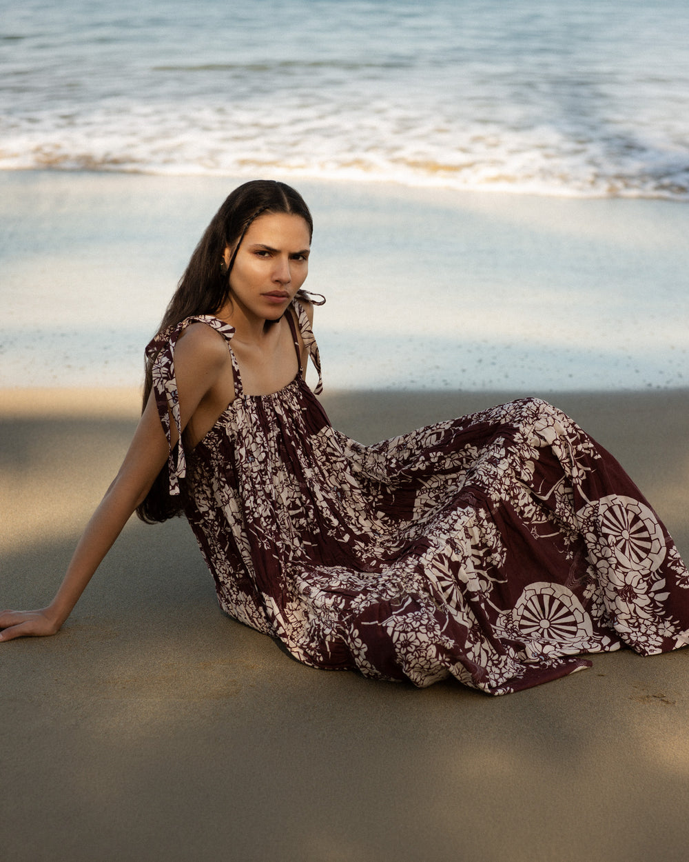Wearing the Emrys Auburn Spoke Dress, a woman sits on the sandy shore by the ocean, gazing at the camera with waves rolling in behind her.