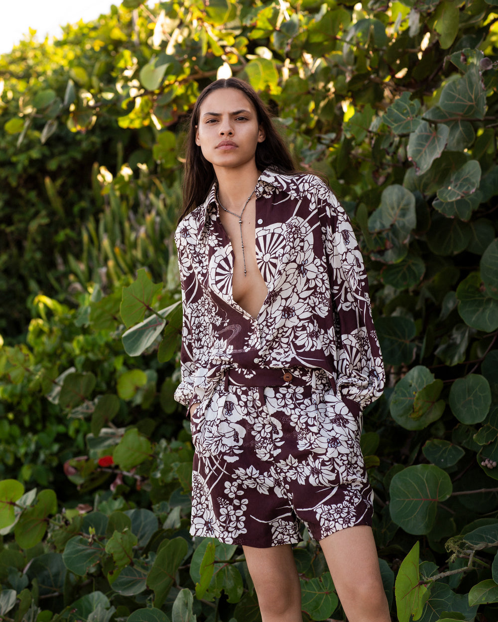 A person stands outdoors in front of leafy green plants, wearing the Deena Auburn Spoke Silk Short and a matching brown-and-white floral shirt set, with the shirt partially unbuttoned.
