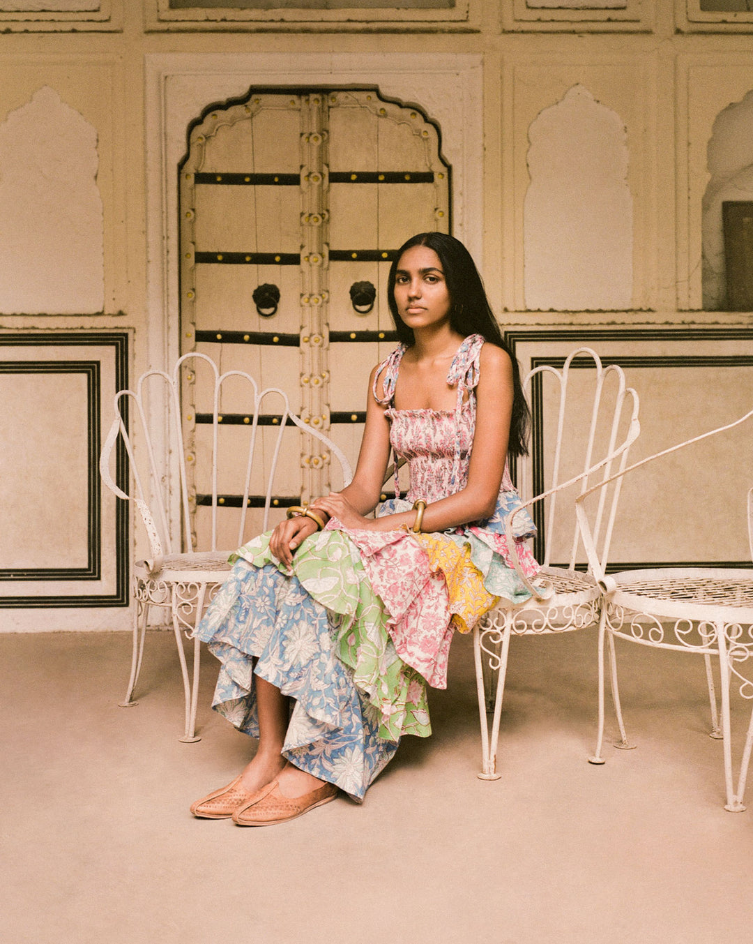 A woman with long hair wears the Club Tropical Ruffle Dress, sitting on a white wrought-iron chair in front of an ornate door. The background showcases intricate details with arches and a frame, enhancing the elegant setting.