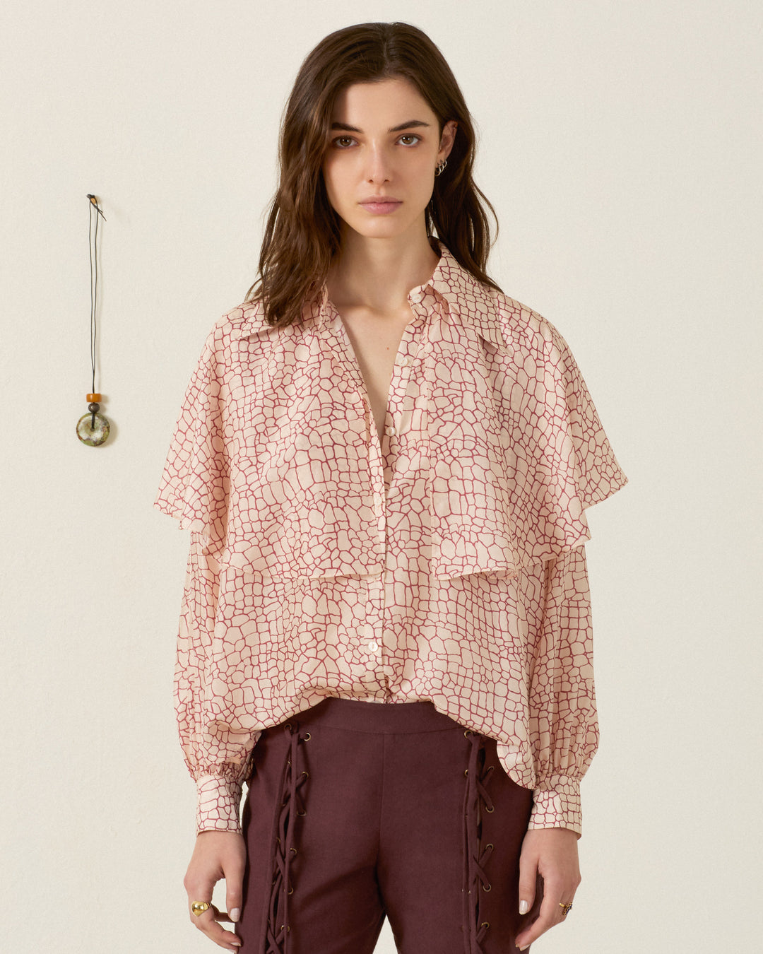 A woman stands indoors facing the camera against a plain wall adorned with a small pendant, wearing the Eleanor Crocodile Silk Shirt— a pink patterned, hand-loomed silk blouse with layered details—paired with maroon pants featuring lace-up sides.
