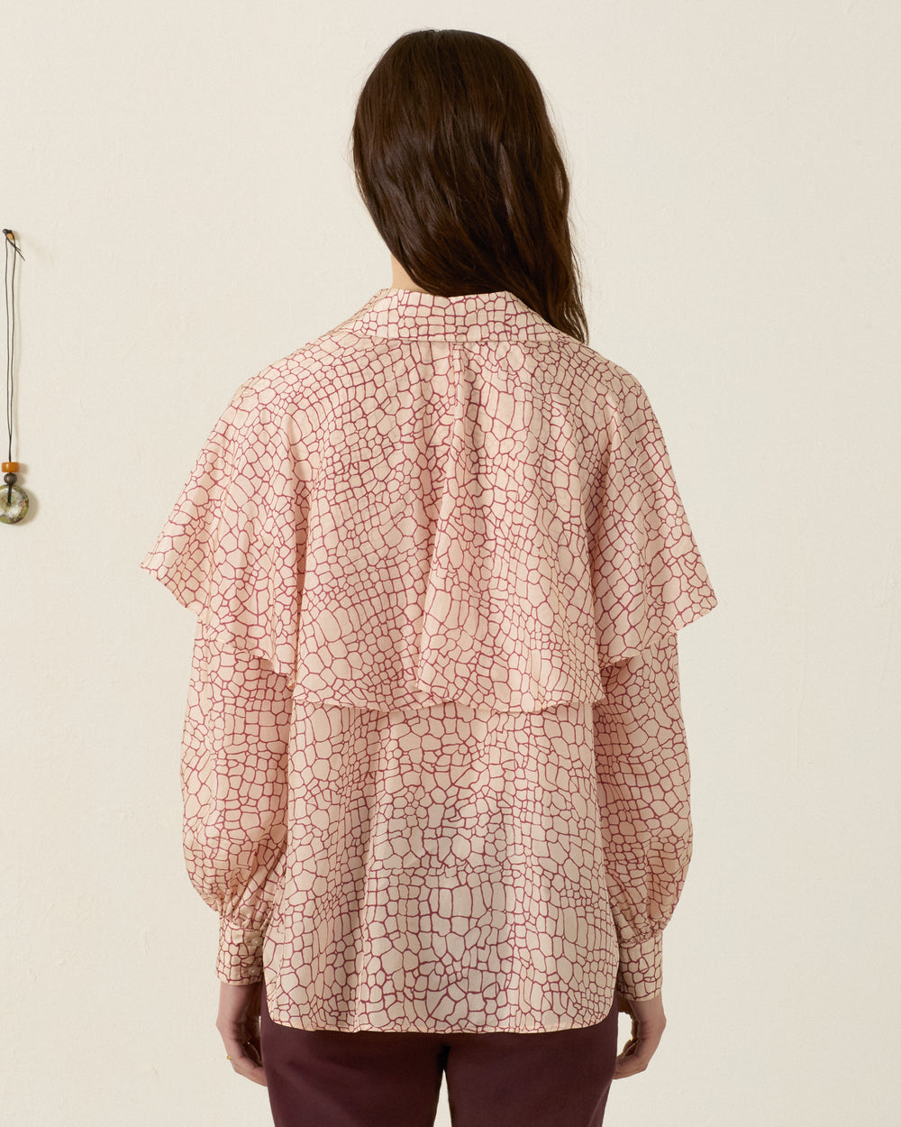 Person facing away, wearing the Eleanor Crocodile Silk Shirt—a long-sleeve, light pink blouse with a subtle crocodile print—and dark pants, standing against a plain light-colored wall.