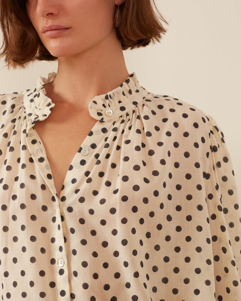 A person is wearing the Annabel Cream Spot Shirt, which features black polka dots and hand-printed tencel ruffled collar details with buttons down the front. They have short brown hair, and the background is a plain light wall.