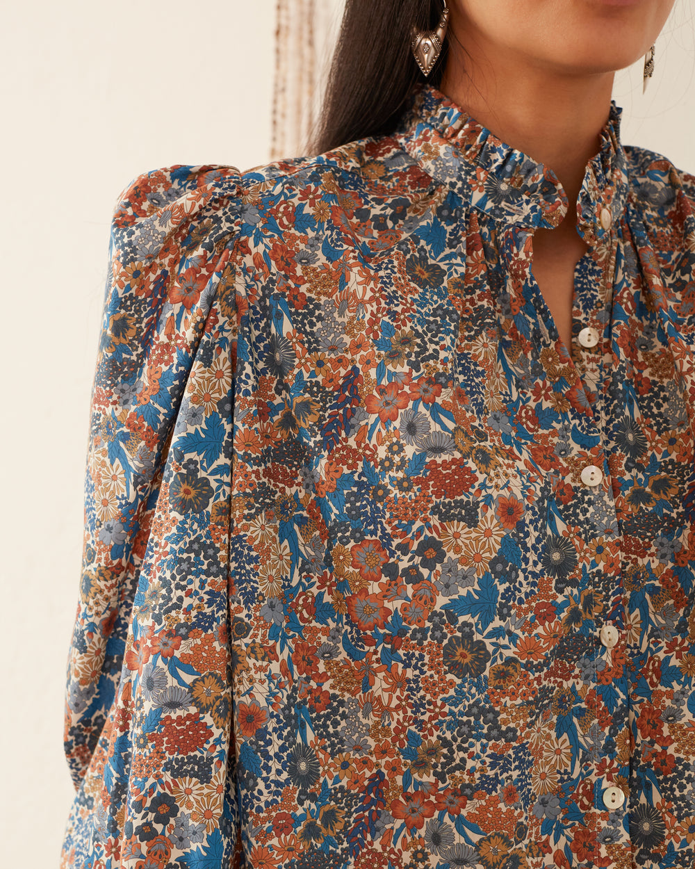 A person wearing the Annabel Liberty Blue Shirt, crafted from Liberty of London's signature cotton lawn and featuring blue, rust, and white floral patterns with a ruffled collar and button-up front. The background is softly blurred.