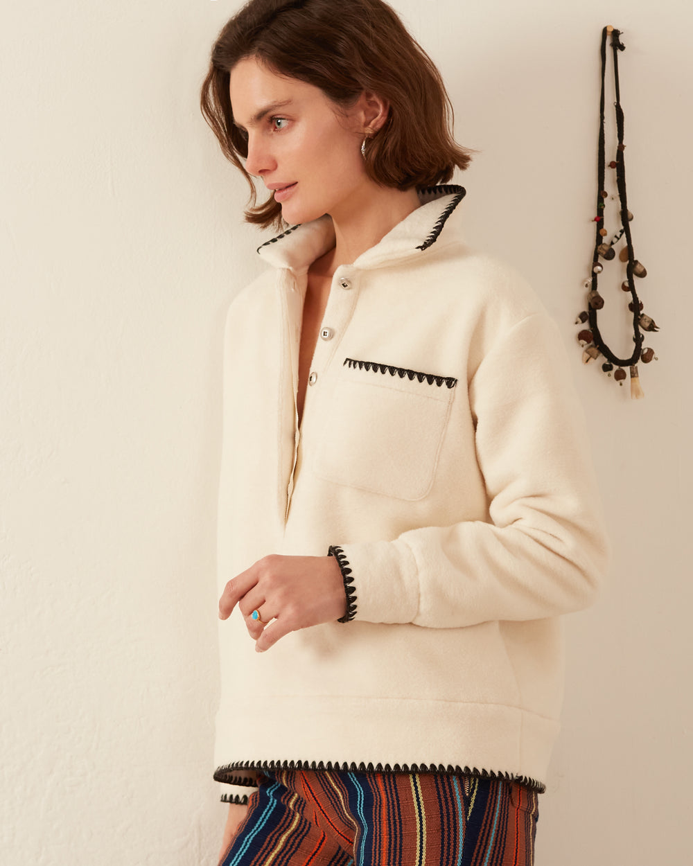 A person wearing the Arden Snow Knit Top, featuring embroidered stitching, stands against a white wall. They have short brown hair and are gazing to the side. A necklace hangs on the wall, and they are wearing colorful striped pants made from Japanese cotton.