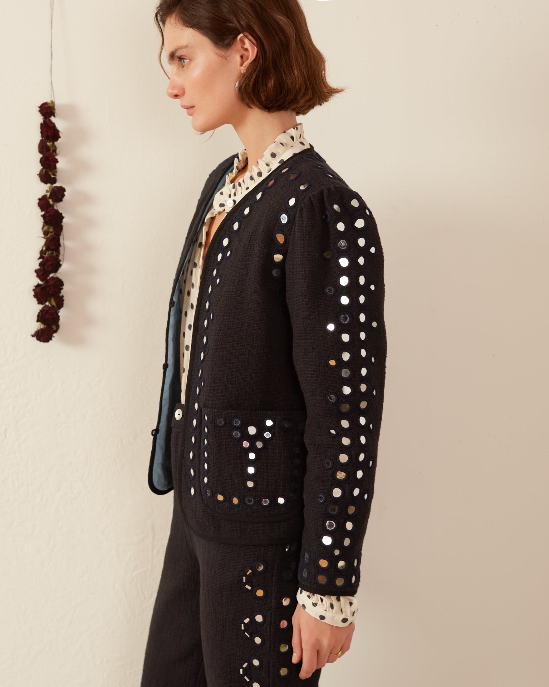 A person with short brown hair is wearing the Billie Midnight Mirror Jacket and pants featuring circular cut-out patterns with hand-embroidered mirrors. They stand in profile against a neutral wall, next to a hanging string of dried flowers.