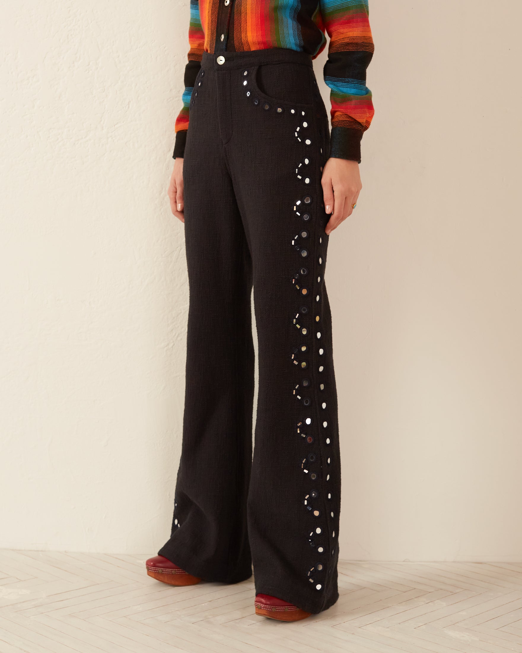 A person wearing the Charlie Black Beauty Pant, featuring decorative buttons and hand-embroidered mirrors, stands against a white wall. Their ensemble is completed by a colorful, striped sweater and red shoes, with only the lower half of their body visible.