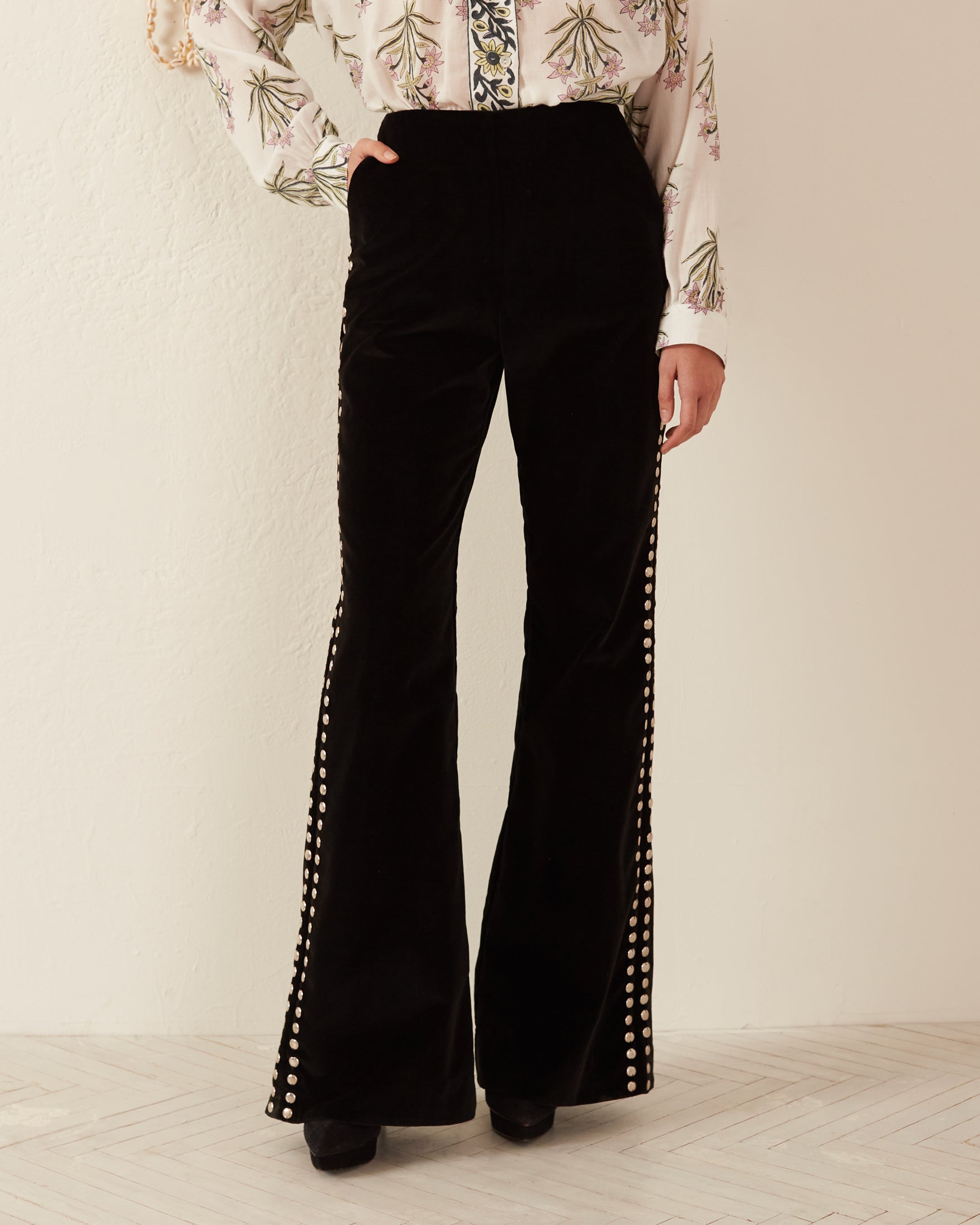 A person wearing the Charlie Rodeo Velvet Pant in black cotton velvet with flared legs and pearl embellishments along the sides, paired with a patterned blouse, presenting a vintage-inspired style.