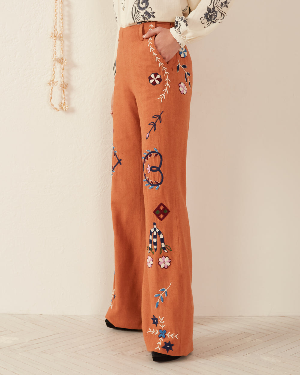 A person dressed in the Charlie Sienna Poppy Embroidered Pant stands against a beige wall. They wear a patterned shirt with one hand on their hip. A string of beads adorns the background, enriching the scene's texture and charm.