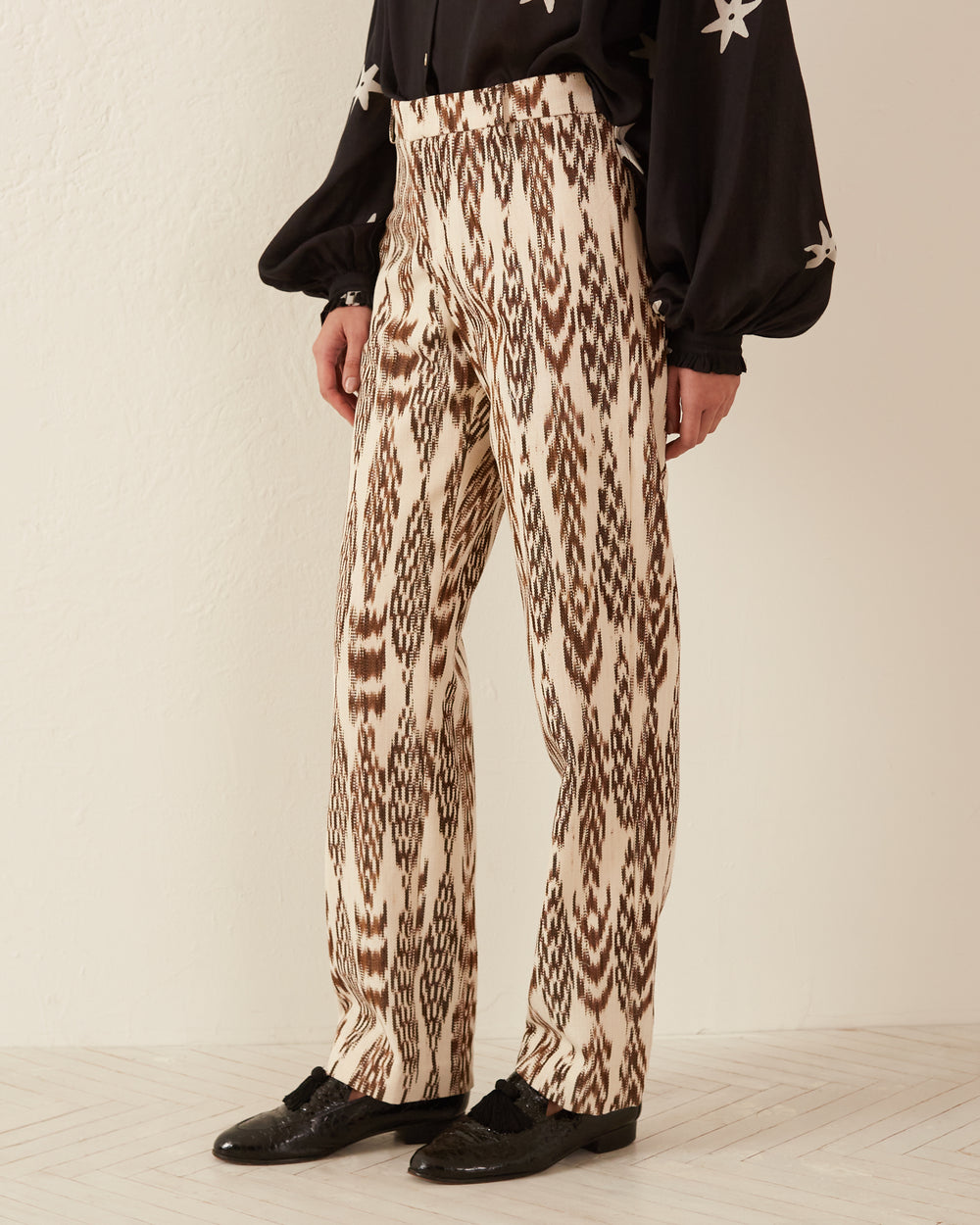 A person wearing Ezra Feather Ikat Pants in cream, showcasing a brown abstract pattern similar to Guatemalan ikat, stands against a light-colored wall. They pair the pants with a black long-sleeve shirt adorned with star designs and black shoes.