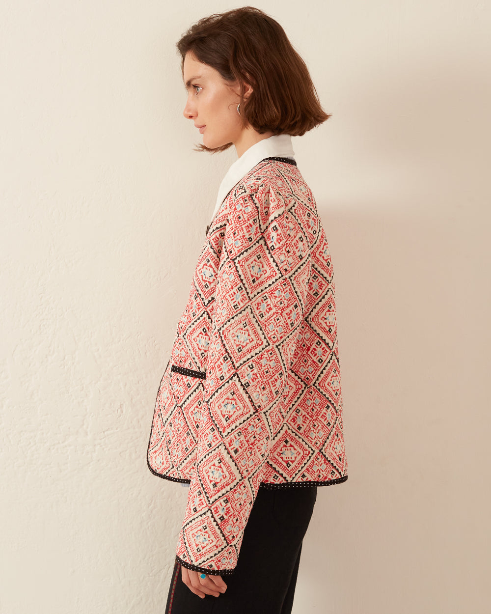 A person, with short brown hair, is shown in side profile wearing a Pasha Jacquard Jacket in a vibrant pink and white pattern adorned with black trim. The jacket showcases a diamond design and is crafted using traditional techniques. They are standing against a plain light-colored wall.