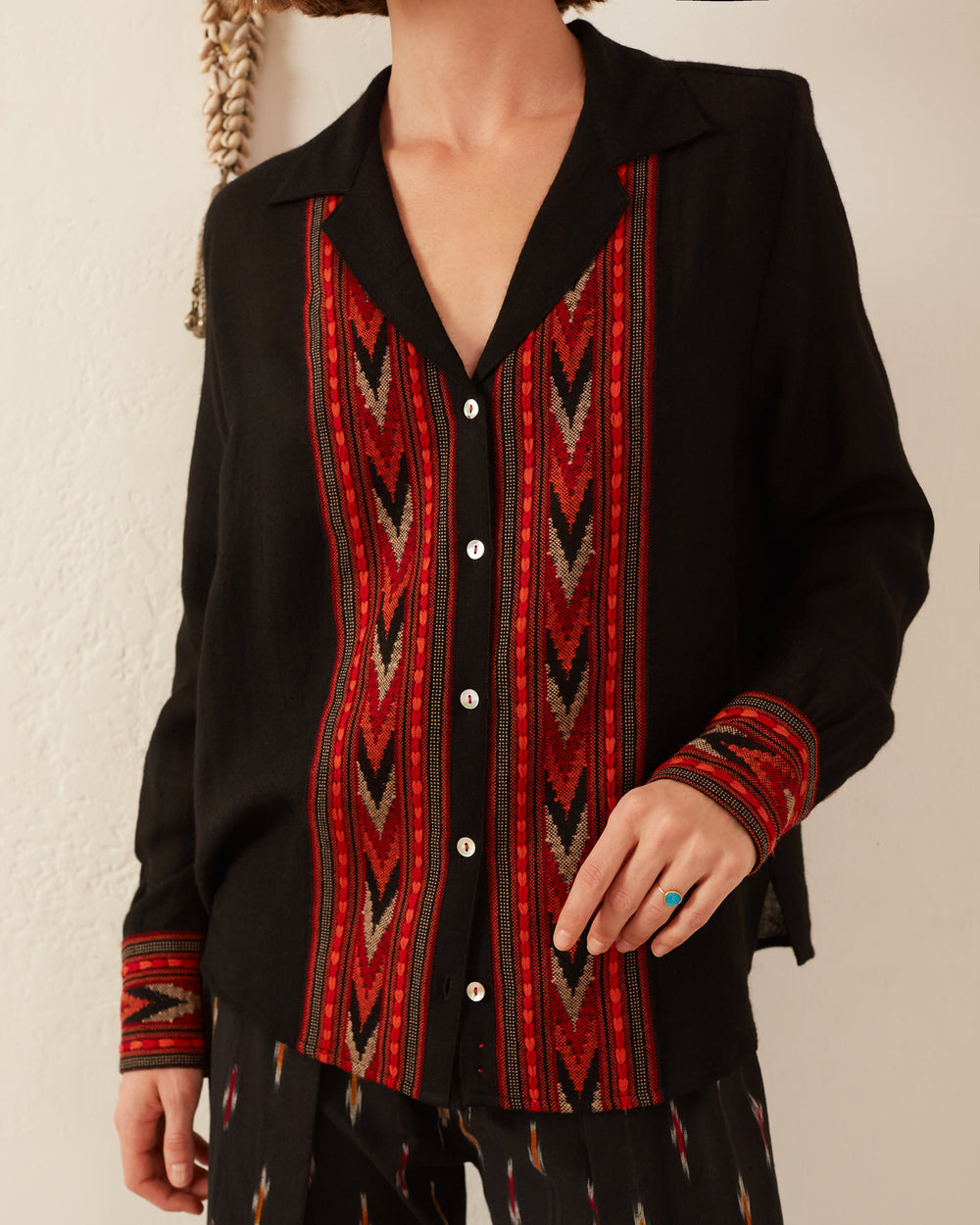 The individual is dressed in a Patti Arrow Shirt, showcasing red and multicolored geometric designs created using traditional techniques on the front and sleeves. This relaxed-fit shirt includes a collared neckline and button-down front. They have a hand resting on their hip, adorned with a turquoise ring.