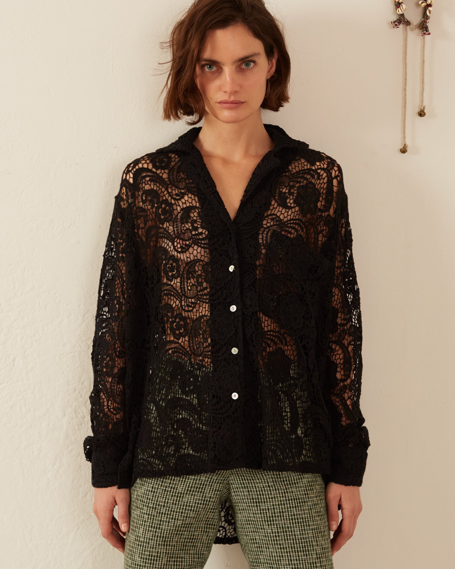 A person with short brown hair stands against a white wall, wearing the Patti Black Floral Lace Shirt over houndstooth pants. A decorative wall hanging is partially visible on the right. The relaxed fit of the outfit adds an effortless touch to their style.