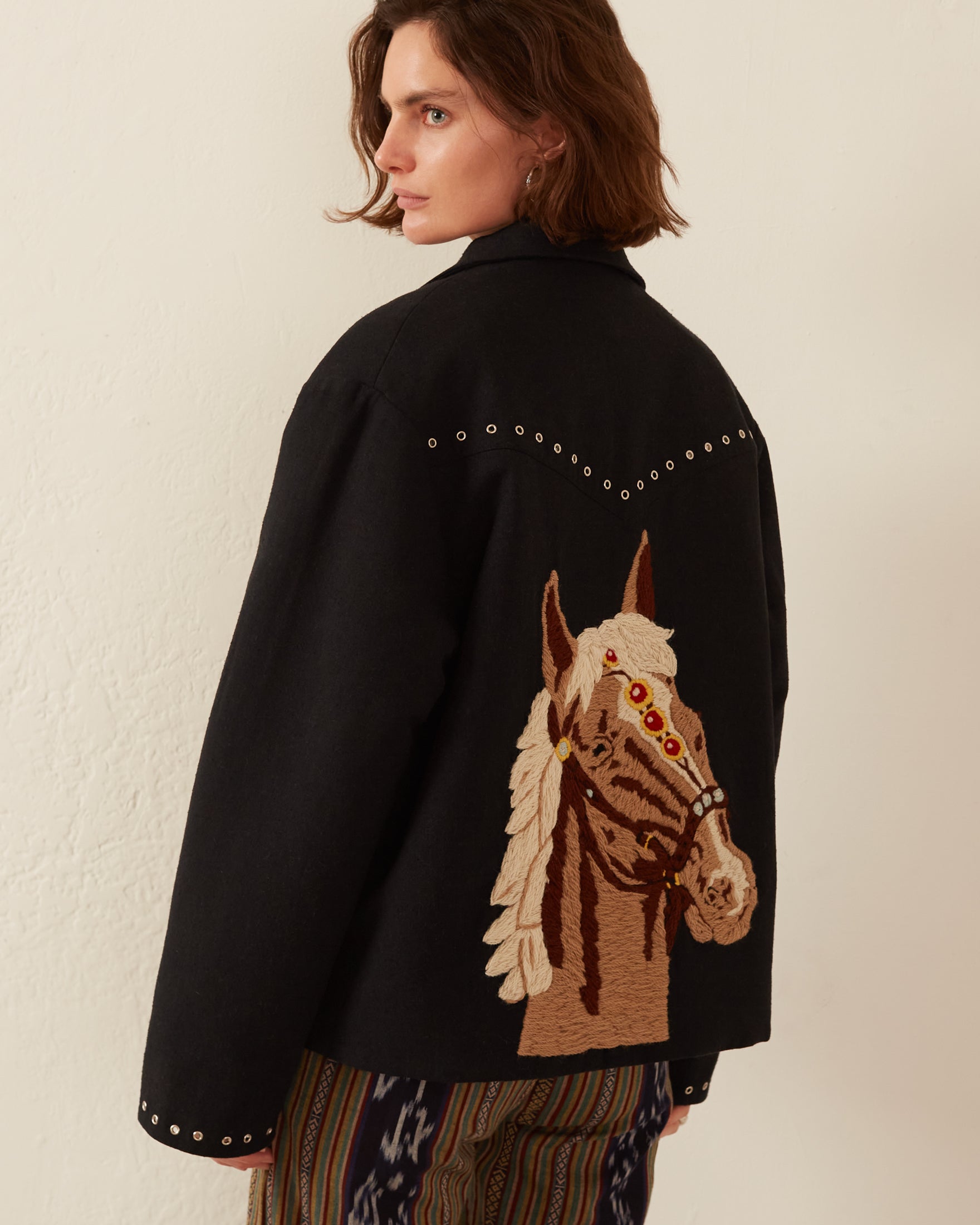 A person with shoulder-length brown hair wears the Western Horse Jacket, showcasing a detailed hand-embroidered horse on the back and adorned with small silver studs on the shoulders. They pose against a plain, light-colored wall.